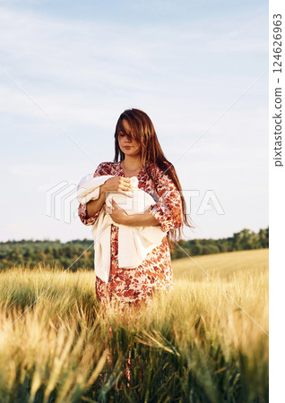 Standing and holding her newborn baby. Gorgeous woman is on the agricultural field 124626963