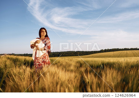 Standing and holding her newborn baby. Gorgeous woman is on the agricultural field 124626964