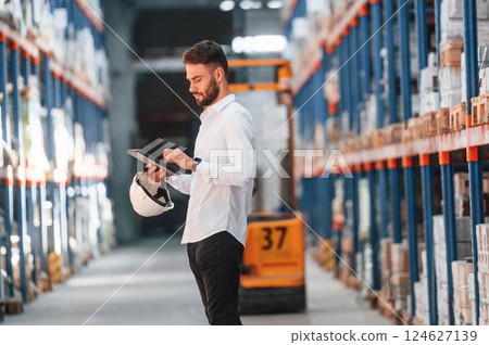 Standing and holding tablet in hands. Storage worker is in the warehouse with bunch of products Standing and holding tablet in hands. Storage worker is in the warehouse with bunch of products 124627139