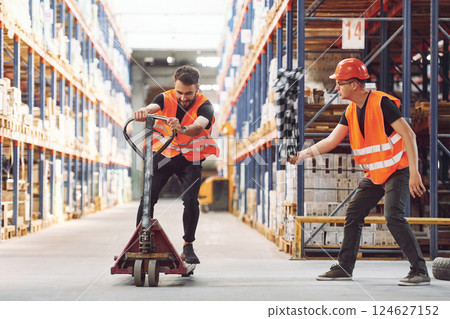 Man riding trolley platform. Having fun. Two storage workers is in the warehouse Man riding trolley platform. Having fun. Two storage workers is in the warehouse 124627152