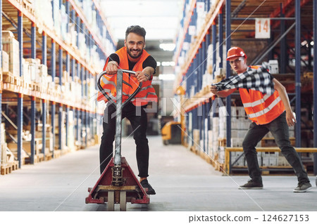 Man riding trolley platform. Having fun. Two storage workers is in the warehouse Man riding trolley platform. Having fun. Two storage workers is in the warehouse 124627153