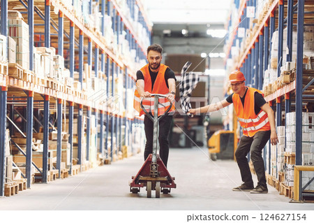 Man riding trolley platform. Having fun. Two storage workers is in the warehouse Man riding trolley platform. Having fun. Two storage workers is in the warehouse 124627154