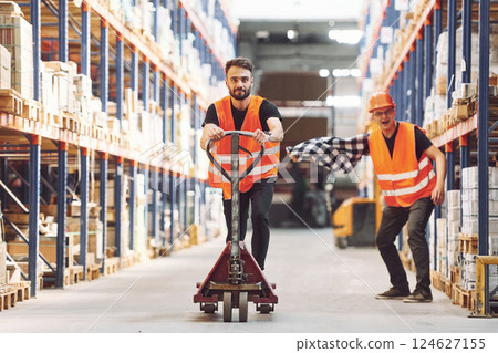 Man riding trolley platform. Having fun. Two storage workers is in the warehouse Man riding trolley platform. Having fun. Two storage workers is in the warehouse 124627155