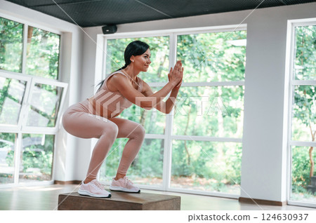 Standing on a cube and doing squats. Woman in sportive clothes is having fitness day in the gym Standing on a cube and doing squats. Woman in sportive clothes is having fitness day in the gym 124630937