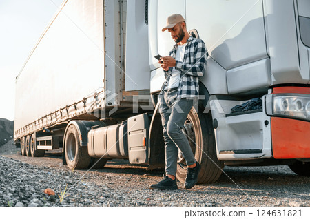 Leaning by the vehicle and using phone. Young truck driver is at daytime Leaning by the vehicle and using phone. Young truck driver is at daytime 124631821