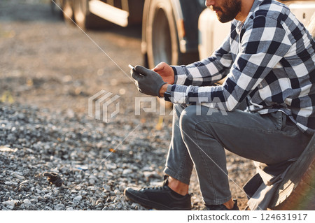 Sitting with phone, waiting for help. Damaged tire. Young truck driver is with his vehicle at daytime Sitting with phone, waiting for help. Damaged tire. Young truck driver is with his vehicle at daytime 124631917