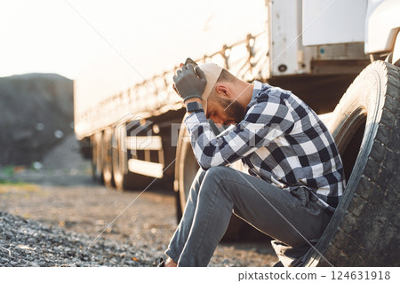 Sitting with phone, waiting for help. Damaged tire. Young truck driver is with his vehicle at daytime 124631918