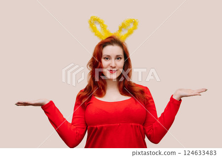 Young red-haired woman in red dress wearing easter costume with bunny ears. Easter concept, copy space. Studio advertising portrait of smiling woman. 124633483