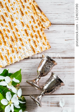 Happy Passover flatlay. Jewish matzo bread and wine glasses on white wooden background. Holiday greeting card with flowers. Copy space. Vertical photo. Happy Passover flatlay. Jewish matzo bread and wine glasses on white wooden background. Holiday greeting card with flowers. Copy space. Vertical photo. 124633490