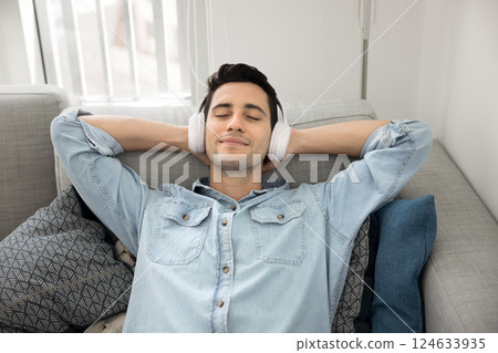 Man lying on sofa wearing headphones listening favourite track Man lying on sofa wearing headphones listening favourite track 124633935