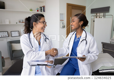 Happy diverse female docs shaking hands in hospital examination room Happy diverse female docs shaking hands in hospital examination room 124634025
