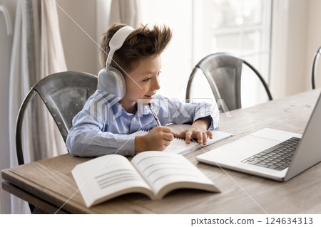 Little boy wearing headphones engaged in learning sits at table Little boy wearing headphones engaged in learning sits at table 124634313