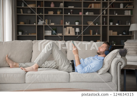 Millennial male relax on sofa with earphones connected to smartphone Millennial male relax on sofa with earphones connected to smartphone 124634315