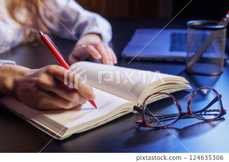 A person writing in a notebook with a red pen, a laptop and a pen holder in the background. Eyeglasses are placed on the table next to the notebook. A person writing in a notebook with a red pen, a laptop and a pen holder in the background. Eyeglasses are placed on the table next to the notebook. 124635306