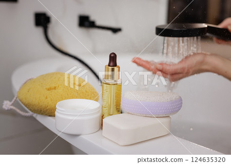 Spa products and skincare essentials on bath edge, hand under shower water in modern bathroom, self-care concept. 124635320