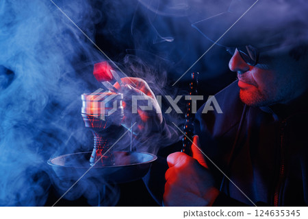 Man smoking hookah, adjusting hot charcoal with tongs in a smoky, moody atmosphere, illuminated by red and blue lighting, Man smoking hookah, adjusting hot charcoal with tongs in a smoky, moody atmosphere, illuminated by red and blue lighting, 124635345