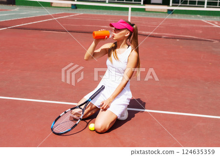Young female tennis player drinking water, holding racket and ball, wearing sportswear and visor, rehydrating after practice on an outdoor court, tennis game 124635359