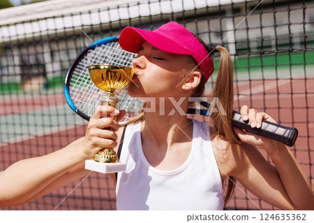 Young female tennis player celebrating victory, kissing trophy on outdoor court. Young female tennis player celebrating victory, kissing trophy on outdoor court. 124635362