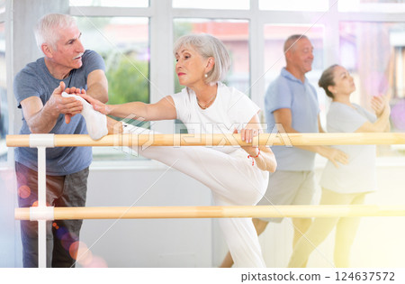 Elderly woman in sportswear, participating in ballet class, mastering developpe movement at barre with aged group and skilled senior male instructor in choreography studio 124637572