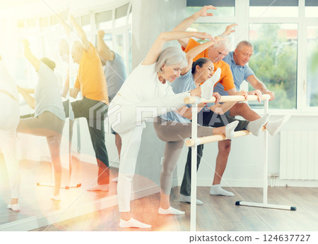 Active mature woman practicing leg stretching holding by ballet barre together with others 124637727