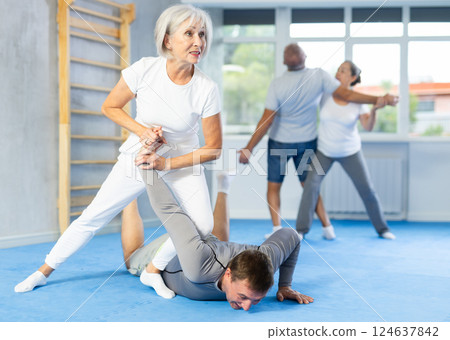 Elderly woman twists arm of attacking man with painful hold in gym. Self-defense lesson Elderly woman twists arm of attacking man with painful hold in gym. Self-defense lesson 124637842