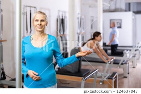 Portrait of smiling aged woman in activewear posing against people exercising on reformer bed machine in Pilates studio Portrait of smiling aged woman in activewear posing against people exercising on reformer bed machine in Pilates studio 124637845