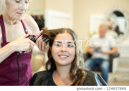 Female hairdresser makes curls the hair of young woman using curling iron 124637849