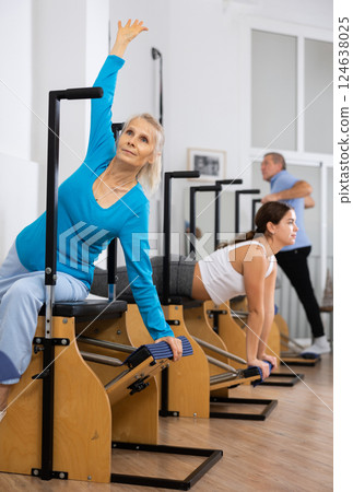 Aged female in activewear concentrating on stretching exercise on Pilates chair equipment in gym. Active and healthy lifestyle rehabilitation concept 124638025