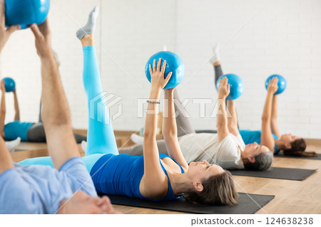 Class of male and women of different ages in active wear pressing balls in hands exercise during Pilates training together in gym Class of male and women of different ages in active wear pressing balls in hands exercise during Pilates training together in gym 124638238