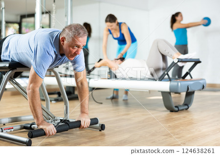 Serious concentrated elderly man performing Pilates exercises on combo chair machine in fitness studio Serious concentrated elderly man performing Pilates exercises on combo chair machine in fitness studio 124638261
