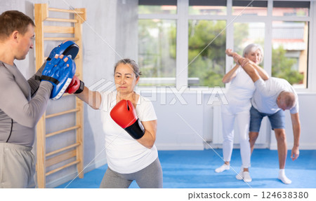 Senior lady and male couch are boxing in gym, man helps student to work out force of blow. Senior lady and male couch are boxing in gym, man helps student to work out force of blow. 124638380
