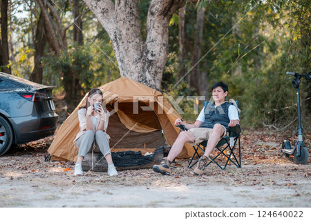 A couple enjoys a peaceful camping trip in the forest, sitting by their tent and car, surrounded by nature. A couple enjoys a peaceful camping trip in the forest, sitting by their tent and car, surrounded by nature. 124640022