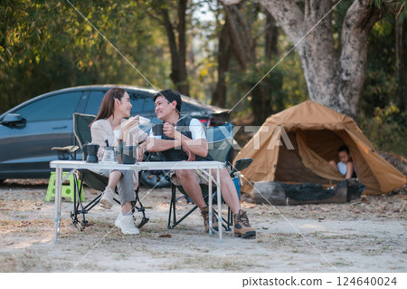 A cheerful couple sitting at a campsite with a tent and car, enjoying nature in a peaceful forest environment. A cheerful couple sitting at a campsite with a tent and car, enjoying nature in a peaceful forest environment. 124640024
