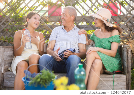 Woman in straw hat is sitting on wooden bench and chatting sweetly with sister and senior father Woman in straw hat is sitting on wooden bench and chatting sweetly with sister and senior father 124642077