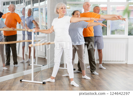 Mature woman in sportswear participating 2nd position of ballet stance at barre with group in choreography studio Mature woman in sportswear participating 2nd position of ballet stance at barre with group in choreography studio 124642297