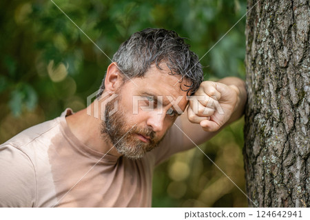 Hispanic mature and brutal bearded man feels tired and sweating after hardworking in the forest leaning on tree outdoor 124642941