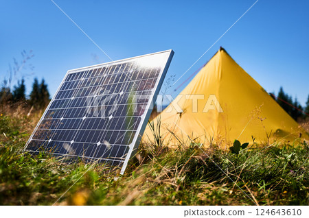 Yellow tent with solar panel set up in grassy field surrounded by dense evergreen trees under clear blue sky. Eco-friendly camping, integration of renewable energy in outdoor activities. 124643610