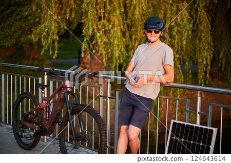 Man cyclist using solar panel for charging electric mountain bike outdoors. Happy guy in helmet, sunglasses leans against railing, holding phone, enjoying peaceful, sunlit setting near lush greenery. 124643614