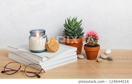 Burning scented candle, books, eyeglasses, and potted plants creating cozy atmosphere Burning scented candle, books, eyeglasses, and potted plants creating cozy atmosphere 124644016