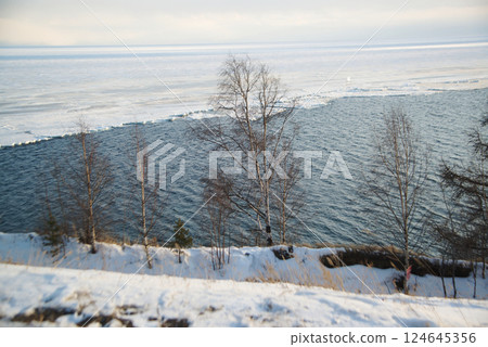 The ice-free Angara River flows out of the frozen Lake Baikal. Melted ice floes on blue water. Snow and dry grass on the shore. Located Baikal lake. The ice-free Angara River flows out of the frozen Lake Baikal. Melted ice floes on blue water. Snow and dry grass on the shore. Located Baikal lake. 124645356