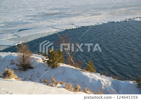 The ice-free Angara River flows out of the frozen Lake Baikal. Melted ice floes on blue water. Snow and dry grass on the shore. Located Baikal lake. 124645359