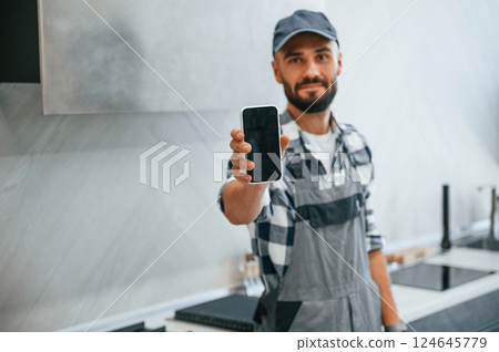 Holding smartphone in hand. Plumber in uniform is indoors at home 124645779