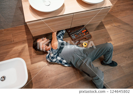 On the floor with case of equipment. Plumber in uniform is indoors at home On the floor with case of equipment. Plumber in uniform is indoors at home 124645802