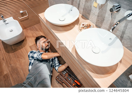 Laying down on the ground, fixing the sink. Plumber in uniform is indoors at home 124645804