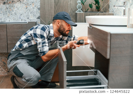 Looking under the sink and using flashlight. Plumber in uniform is indoors at home 124645822
