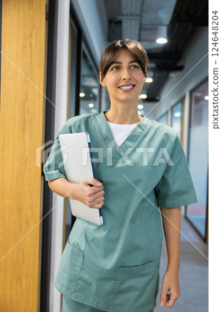Female physician in medical uniform standing in hospital corridor holding laptop in hands Female physician in medical uniform standing in hospital corridor holding laptop in hands 124648204