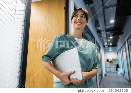 Female physician in medical uniform standing in hospital corridor holding laptop in hands Female physician in medical uniform standing in hospital corridor holding laptop in hands 124648208