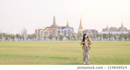 Traveler asian woman in her 30s, backpack slung over her shoulder, explores Wat Pra Kaew with childlike wonder. Sunlight dances on the golden rooftops, and a carefree smile Traveler asian woman in her 30s, backpack slung over her shoulder, explores Wat Pra Kaew with childlike wonder. Sunlight dances on the golden rooftops, and a carefree smile 124648924