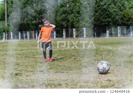 Little boy in orange t-shirt stands on grassy field in front of soccer ball, seen through goal net. Little boy in orange t-shirt stands on grassy field in front of soccer ball, seen through goal net. 124649416
