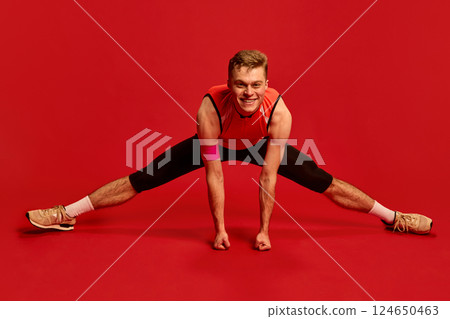 Happy athletic man doing wide-legged stretch on floor, arms pressing down, smiling during intense flexibility workout. Happy athletic man doing wide-legged stretch on floor, arms pressing down, smiling during intense flexibility workout. 124650463